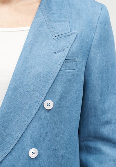 Blazer bleu clair avec revers cranté, deux boutons blancs et poche passepoilée porté sur un haut côtelé blanc.