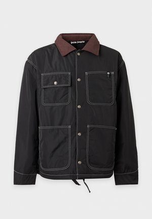 Homme portant une casquette noire, une veste noire avec des coutures blanches, un sweat à capuche rouge, un pantalon beige tie-dye et des baskets noires, debout face à l'avant.
