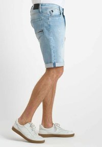 Shorts de mezclilla azul claro con dobladillo enrollado, que presentan detalles desgastados y un bolsillo trasero, combinados con zapatillas blancas y un diseño de cordones plano.