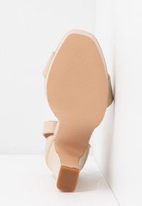 Sandale beige à talons hauts montrant la semelle avec une zone des orteils texturée et un talon bloc, positionnée en position verticale contre un fond blanc d’angle.