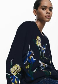 Navy knitted sweater featuring colorful floral embroidery in yellow, blue, and green. Loose fit with ribbed cuffs and hem.