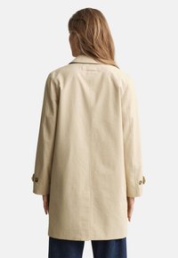 Abrigo tipo trench color beige con corte recto, cuello y botones visibles en las mangas; hecho de tela texturizada, que llega hasta la mitad del muslo.