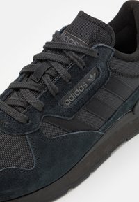 Sneaker Adidas neri caratterizzati da un mix di materiali scamosciati e in mesh, con un design testurizzato, lacci tondi e suola in gomma.