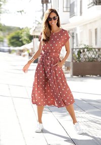Bruine jurk met polka dots, voorzien van een strikceintuur en zakken, kniehoogte. G gedragen met witte sneakers, tegen een zonovergoten buitenachtergrond.