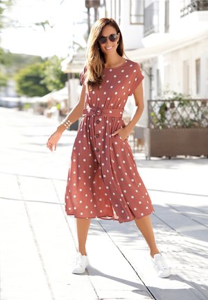 Bruine jurk met polka dots, voorzien van een strikceintuur en zakken, kniehoogte. G gedragen met witte sneakers, tegen een zonovergoten buitenachtergrond.