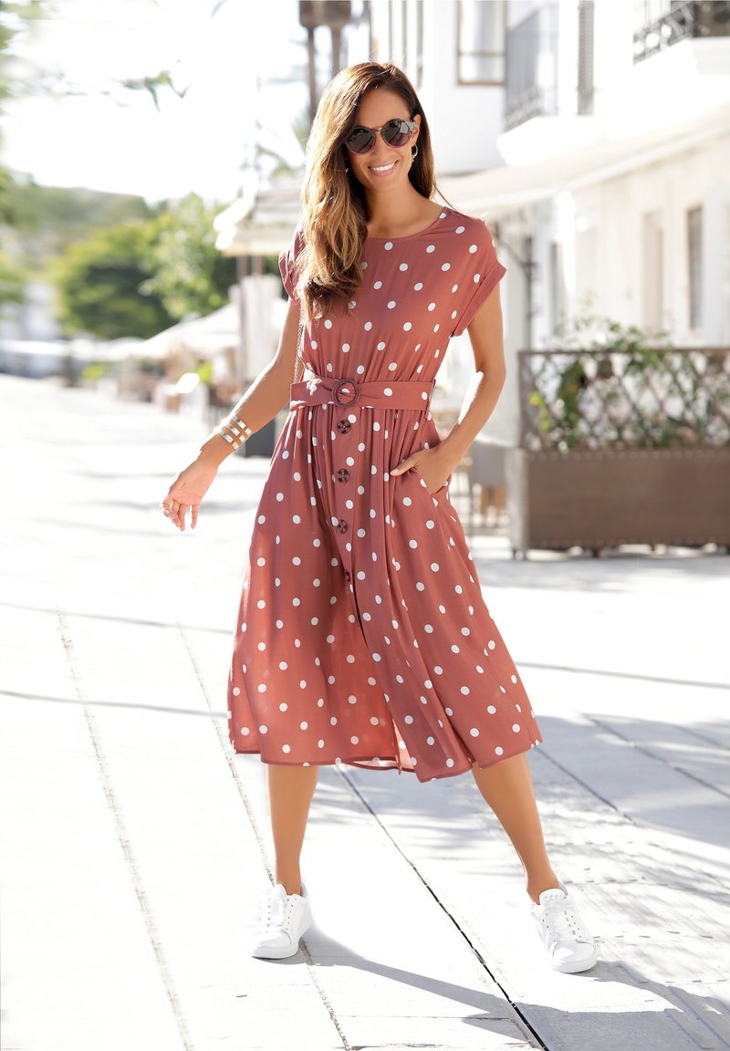 Bruine jurk met polka dots, voorzien van een strikceintuur en zakken, kniehoogte. G gedragen met witte sneakers, tegen een zonovergoten buitenachtergrond.