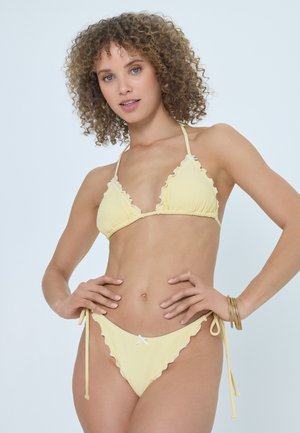 Young woman with curly hair wearing a light yellow ruffled bikini and gold bangles, standing with hands on hips against white background.