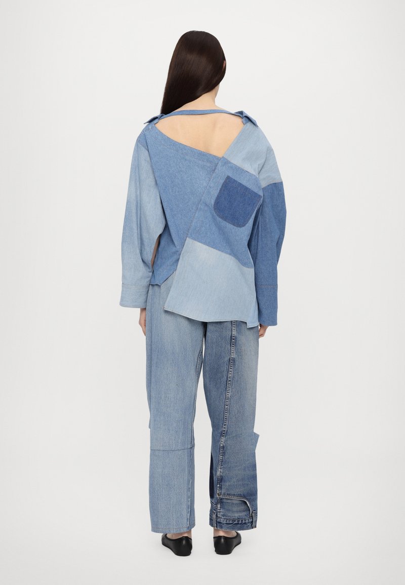 Denim shirt with varying blue shades, asymmetric design, open back, and long sleeves. Paired with loose blue jeans. Flat black shoes.