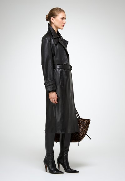 Donna in profilo che indossa un trench coat in pelle nera con cintura, stivali neri con tacco fino al ginocchio, mentre tiene una grande borsa a tracolla con stampa leopardata.