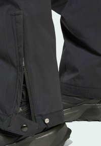 Calças impermeáveis pretas com fechos de correr e botões nos punhos. Tecido texturizado com um acabamento mate, combinado com umas robustas botas de caminhada pretas.