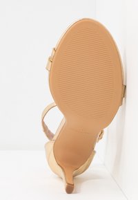 Semelle d'une sandale dorée à talons avec brides, semelle extérieure couleur tabac texturée, petit talon, inclinée contre un fond blanc en angle.