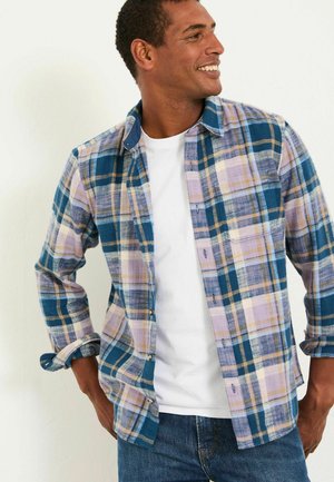 Karierte Flanellbluse in Blau, Lila und Beige; mit Knopfleiste, Brusttasche und hochgekrempelten Ärmeln; über einem weißen T-Shirt getragen.