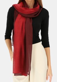 Czerwona szalik o gradientowym przejściu kolorów, wykonany z lekkiego materiału, prezentujący gładką fakturę. Elegancko opada wokół szyi, uzupełniając czarną bluzkę i neutralne spodnie.