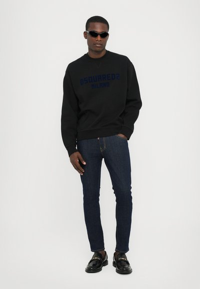 Sweatshirt noir avec le texte bleu "DSQUARED2 MILANO", jean sombre et mocassins noirs brillants. Le mannequin porte une tenue ajustée et des lunettes de soleil.