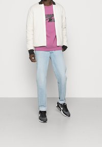 Uomo in giacca bomber bianca, felpa grafica viola, jeans chiari con risvolti e sneaker nere Nike, in piedi contro un muro semplice.