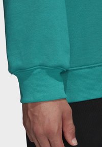Teal-coloured huppari hihassa on joustinta, ja kyynärpäässä on sauma ilman saumoja. Kankaan pinta on sileä. Käsi näkyy reunan kohdalla.