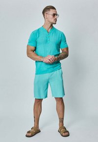 Chemise henley turquoise à manches courtes avec patte de boutonnage, assortie à un short assorti. Portée avec des sandales marron, présentant un tissu texturé.