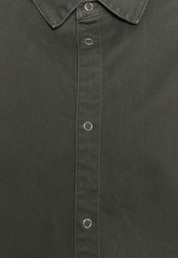 Tummanvihreä puuvillainen nappipaita, jossa on terävä kaulus ja pyöreät hopeiset metalliosat paidan keskiosassa.