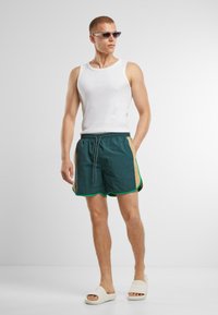 Zöld fürdőnadrág sárga kiegészítőkkel és zöld szegéllyel, fehér tank top-pal és világos bézs papuccsal párosítva. Átlátszó napszemüveg viselve.
