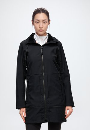 Une femme porte un long manteau noir zippé sur un haut blanc à col montant, sur un fond uni et clair.