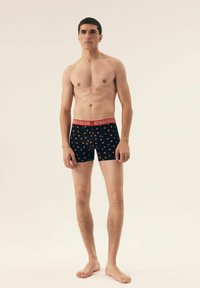 Czarne slipki z czerwonym paskiem w talii, z wzorem małych czerwonych serc. Gładki materiał z dopasowanym krojem. Model stoi boso.