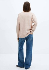 Persoon met schouderlang bruin haar, gekleed in een losse beige trui en wijde blauwe jeans, staand met één hand in de zak, met de rug naar de camera.