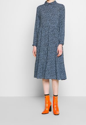 Personne portant une robe midi à manches longues bleu marine à fleurs, avec des bottines à talons hauts orange vif, debout sur un sol gris.