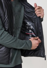 Crna puffer prsluk s glatkom vanjštinom, s patentnim zatvaračem sprijeda i džepom s ikonama i tekstom na unutarnjoj podstavi. Nositi preko teksturirane zelene majice.
