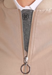 Chaqueta bomber beige con un zipper plateado y un tirador redondo. El interior incluye un forro de fieltro gris, con un cuello estructurado y tejido texturizado.