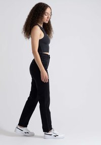 Jeans pretos de cintura alta combinados com uma camisola justa preta e ténis brancos com detalhes pretos, vista lateral que destaca a silhueta e o ajuste.