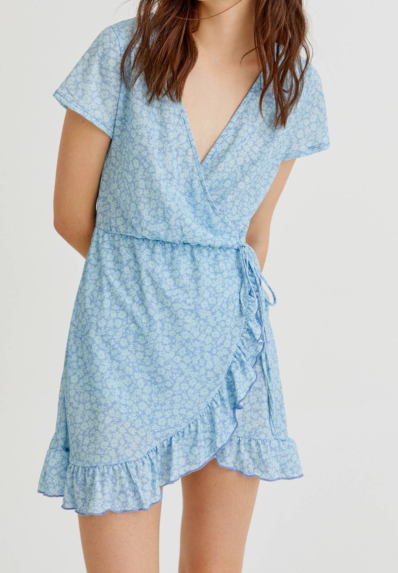 Femme portant une robe portefeuille à manches courtes, bleue claire à fleurs, avec un ourlet volanté et un lien sur le côté, sur un fond uni.