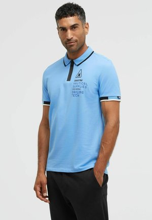 Man wearing light blue polo shirt with nautical text and black pants, standing with one hand in pocket against plain background.