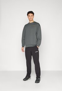 Sweat-shirt en velours texturé vert foncé à manches longues, associé à un pantalon de sport noir avec un logo blanc et des chaussures de sport noires.