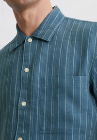Blau gestreiftes Hemd aus leichtem Stoff. Verfügt über einen Kragen, eine Brusttasche und dezente weiße Streifen auf einem einfarbigen Hintergrund. Vier sichtbare Knöpfe.