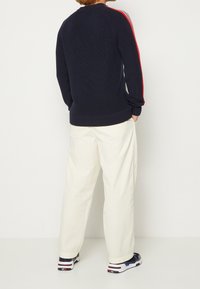 Marineblauer Strickpullover mit roten und weißen Streifen an den Ärmeln, gerippten Bündchen und Saum, kombiniert mit weißen Hosen und marineblauen Sneakers.