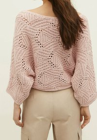 Frau mit langen braunen Haaren, die einen hellrosa gemusterten Strickpullover mit weiten Ärmeln und beigen Hosen trägt, von hinten gesehen.