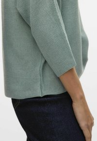 Pull côtelé vert clair à manches trois quarts, avec des fentes sur les côtés à l'ourlet. Assorti avec un jean en denim foncé.