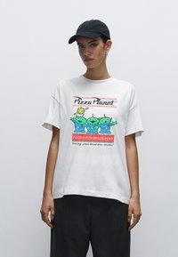 Bílá tričko s barevnými kreslenými mimozemšťany a textem "Pizza Planet" s červenými akcenty, volný střih, kulatý výstřih, krátké rukávy.