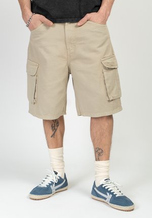 Man wearing beige cargo shorts with side pockets, white crew socks, blue sneakers with white laces, and tattoos on both lower legs.