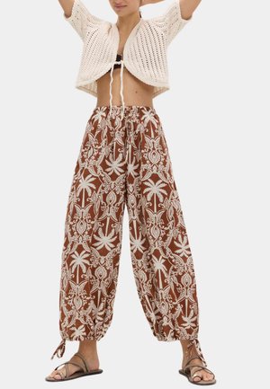 Frau trägt braune, weit geschnittene Hose mit weißem tropischem Druck, weißes gestricktes kurzes Top und braune Riemchensandalen, steht mit erhobenen Armen.