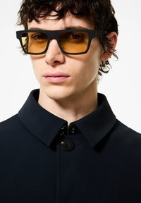 Occhiali da sole neri con lenti arancioni, caratterizzati da una forma quadrata e una montatura spessa, abbinati a un cappotto scuro con colletto e dettaglio a bottoni.