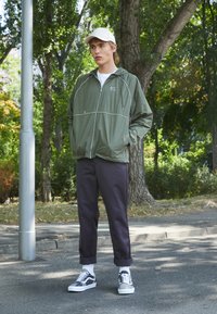Light olive green windbreaker with white piping, black trousers, and patterned black-and-white sneakers. Set in an outdoor urban environment.