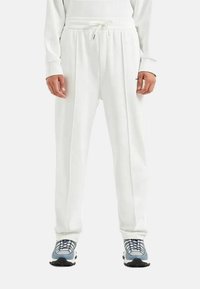 Pantaloni jogger bianchi con vita regolabile, in tessuto morbido, vestibilità ampia, con pieghe laterali e una texture liscia. Abbinati a scarpe da ginnastica grigie.