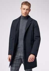 Cappotto trench blu navy con colletto strutturato, due tasche frontali e chiusura a bottoni, indossato sopra un dolcevita grigio e pantaloni a quadri.