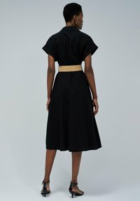 Schwarzes Kleid mit kurzen umgeschlagenen Ärmeln, betonter Taille mit einem Strohbelt, fließendem Rock, aus strapazierfähigem Stoff, Saum fällt unter das Knie.