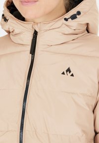 Beige polstret jakke med høj krave, udstyret med en sort lynlås og logo-patch. Stoffet har en glat tekstur med vandret syninger.