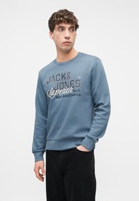 Νεαρός άνδρας που φοράει μια μπλε μπλούζα Jack & Jones με κείμενο, με το ένα χέρι στην τσέπη, κοιτάζοντας προς τα πλάγια σε απλό ανοιχτό φόντο.