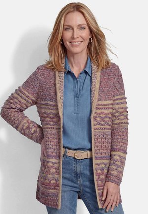 Frau mit schulterlangem blondem Haar, die eine gemusterte Strickjacke über einem blauen Jeanshemd und Jeans trägt, lächelt mit einer Hand in der Hüfte.