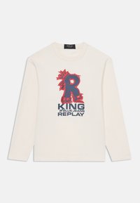 Pitkähihaisen kermaisen puuvillapaidan etuosassa on rohkea punainen ja laivastonsininen graafinen kuvio, jossa lukee "KING of BLUE JEANS REPLAY" sekä tyylitelty kirjain "R."