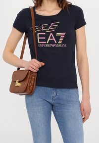 Femme portant un t-shirt bleu marine Emporio Armani avec un logo, un jean bleu clair, et portant un sac à bandoulière en cuir marron avec une fermeture dorée.